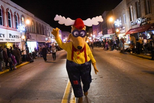 A costumed character resembling a reindeer waves to the crowd during a nighttime parade on a festive street.