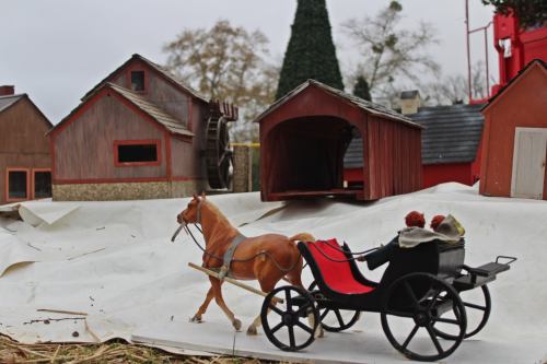 A toy horse pulling a cart with two figures, set against a backdrop of miniature buildings and a cloudy sky.