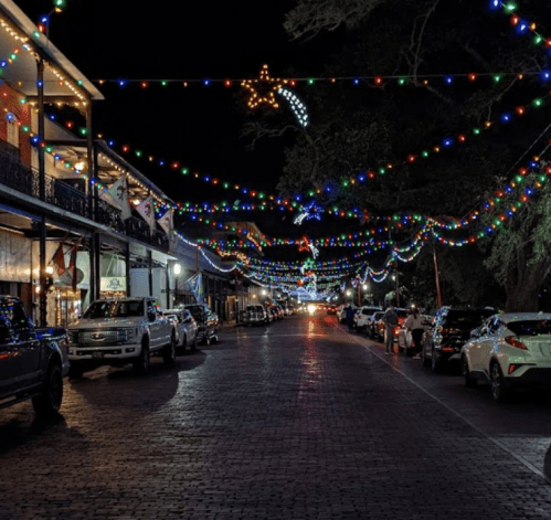 A festive street at night, adorned with colorful lights and decorations, lined with parked cars and historic buildings.