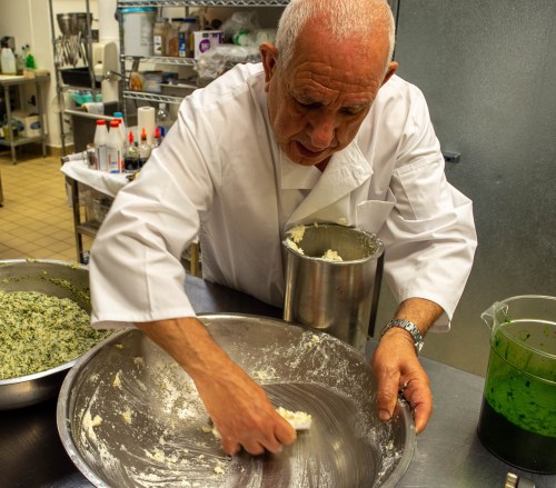 A chef in a white coat mixes ingredients in a large metal bowl in a busy kitchen.