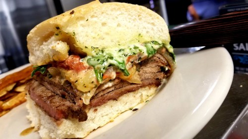 A close-up of a hearty sandwich with layers of beef, greens, and sauce, served on a soft bun.