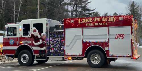 A festive fire truck decorated with lights, featuring Santa Claus waving from the side in a snowy setting.