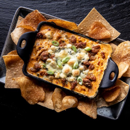 A skillet of cheesy dip topped with green onions, surrounded by crispy tortilla chips on a black plate.