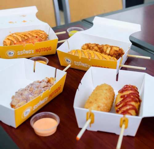 Four hot dogs in takeout boxes, two with sauces, served with dipping sauce on a wooden table.