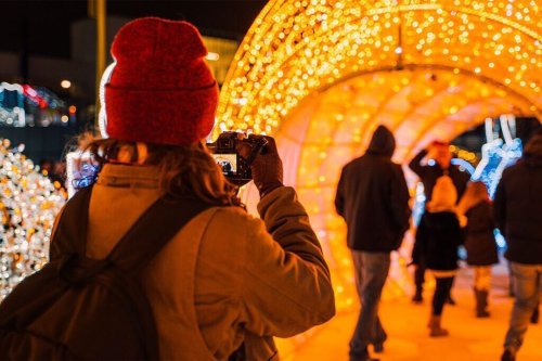 A person in a red hat takes photos of a festive light display while others walk through the illuminated archway.