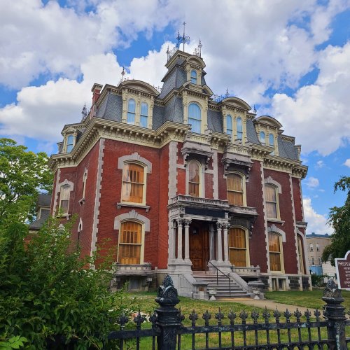 Victorian-style mansion with red brick, ornate details, and large windows, surrounded by greenery under a cloudy sky.