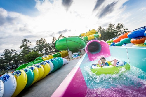 A child slides down a colorful water slide, splashing into a pool, surrounded by inflatable rings and sunny skies.