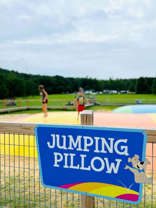 A sign reading "Jumping Pillow" in front of a colorful inflatable jumping area with people in the background.