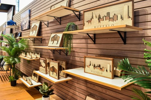 Wooden art pieces depicting cityscapes and buffalo, displayed on shelves against a wooden wall with plants.