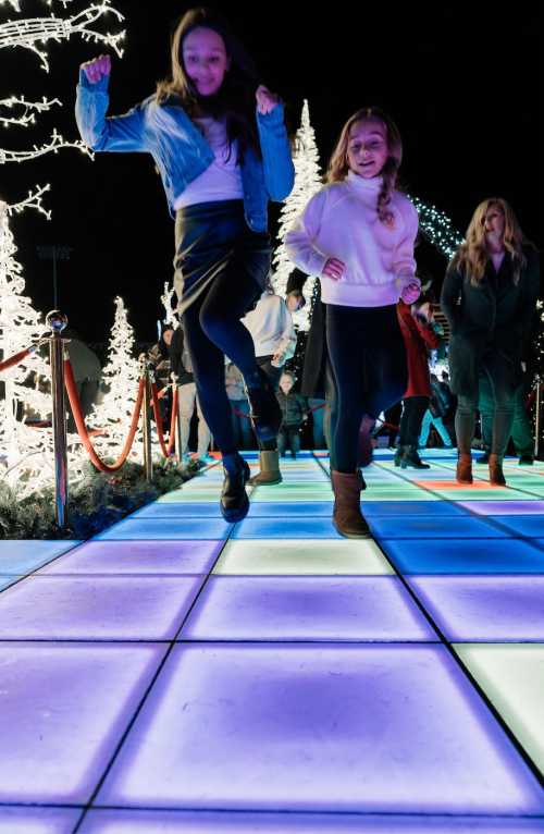 Two girls joyfully dance on a colorful illuminated floor, surrounded by festive lights and holiday decorations.