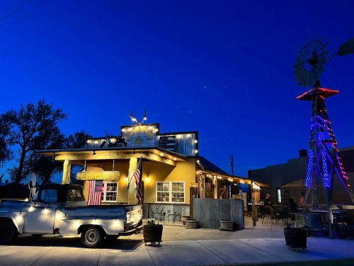 A rustic building with a vintage truck and windmill, illuminated at dusk with blue sky in the background.