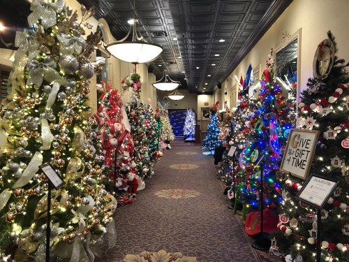 A festive hallway lined with decorated Christmas trees, twinkling lights, and holiday decorations on display.
