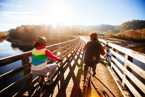Two cyclists ride on a wooden bridge over a river, surrounded by autumn trees and bright sunlight.