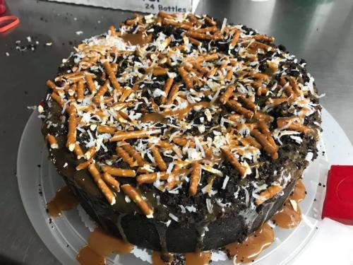 A round chocolate cake topped with caramel, shredded coconut, and pretzel pieces, sitting on a white plate.