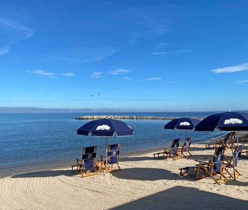 Beach scene with blue umbrellas and chairs on sandy shore, calm water, and a clear blue sky.