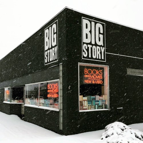 A snowy scene of a black building with large "BIG STORY" signs, featuring windows displaying books and movies for sale.