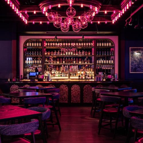 A stylish bar with a vibrant pink-lit atmosphere, featuring a well-stocked shelf of drinks and cozy seating.
