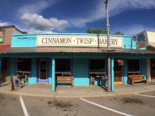 A colorful bakery storefront with a sign reading "Cinnamon Twisp Bakery," featuring benches outside and a clear blue sky.