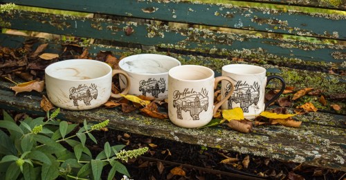 Four ceramic mugs with a design of Poughkeepsie, NY, arranged on a weathered green bench surrounded by autumn leaves.