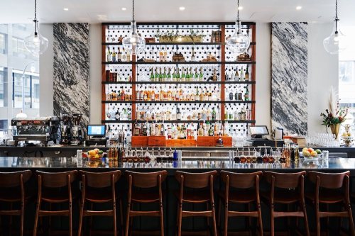 A stylish bar with a marble backdrop, featuring a well-stocked shelf of various bottles and a sleek counter with seating.