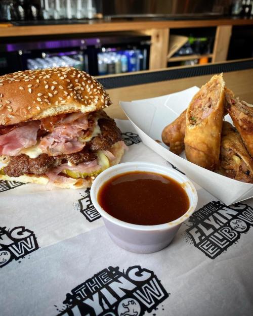 A hearty burger with layers of meat and toppings, served with crispy spring rolls and a side of dipping sauce.