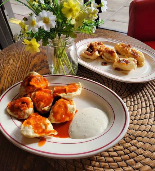 A plate of dumplings drizzled with sauce and a side of dipping sauce, alongside a plate of similar dumplings and flowers.