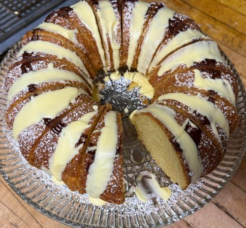 A round bundt cake drizzled with chocolate and white icing, placed on a glass cake stand.