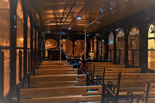 Interior of a vintage trolley with empty wooden benches and large windows, softly lit at night.