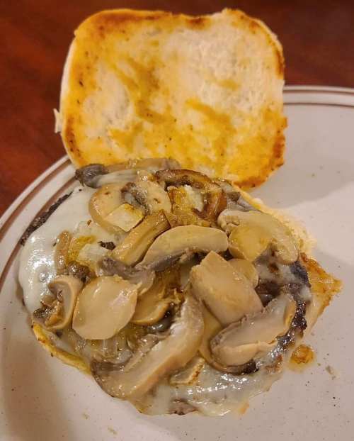 A close-up of a burger with melted cheese and sautéed mushrooms on a toasted bun, partially open on a plate.