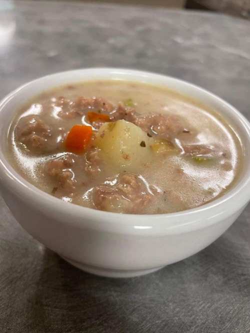 A close-up of a white bowl filled with hearty soup containing meat, vegetables, and potatoes.