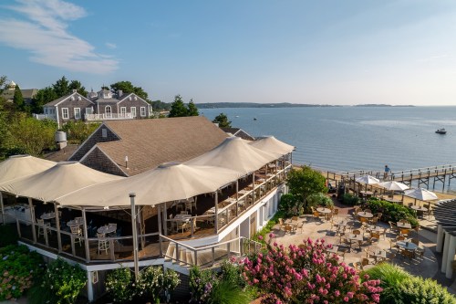 A seaside restaurant with a large patio, surrounded by flowers, overlooking calm waters and distant boats.