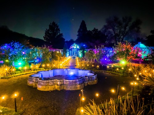 A beautifully lit garden at night, featuring colorful lights, a central fountain, and illuminated pathways.