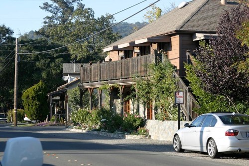 A charming two-story building with a wooden balcony, surrounded by greenery, along a quiet road. A parked car is visible.