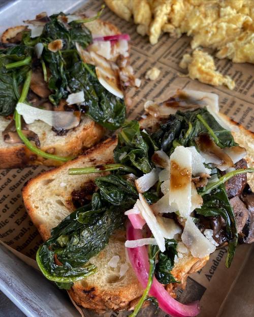 Two slices of toasted bread topped with sautéed spinach, mushrooms, pickled onions, and shaved cheese on a tray.