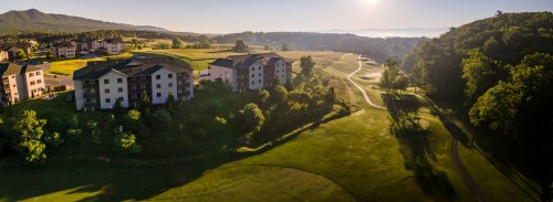 A scenic view of a golf course with buildings and trees, bathed in morning light and misty hills in the background.