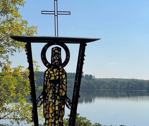 Stained glass figure with a cross, overlooking a serene lake and trees under a clear blue sky.