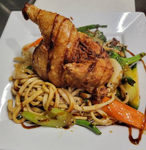 A plate of fried chicken atop noodles and mixed vegetables, drizzled with sauce and garnished with sesame seeds.