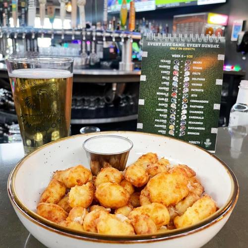 A bowl of golden fried snacks with a dipping sauce, alongside a pint of beer and a game schedule on the bar.