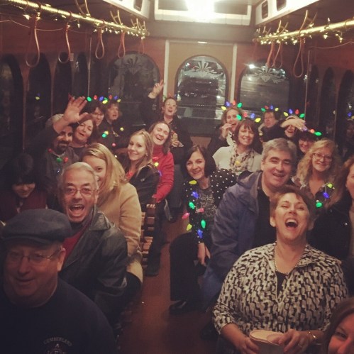 A festive group of people smiling and celebrating inside a decorated trolley, with colorful lights and cheerful expressions.