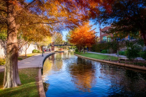 A serene canal lined with trees displaying autumn colors, reflecting in the water under a clear blue sky.