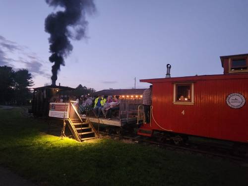 A vintage train with a red caboose and steam rising, passengers seated on a platform at dusk.