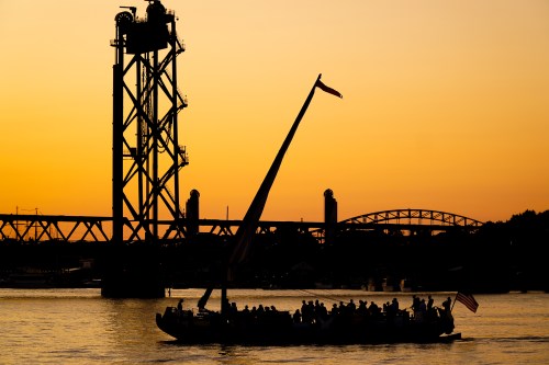 Silhouette of a boat with a sail and passengers against a vibrant sunset, with a bridge and industrial structure in the background.