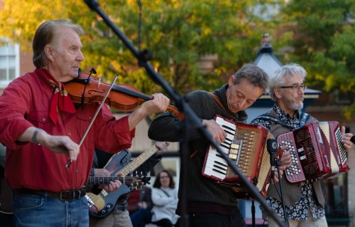 Three musicians perform outdoors, playing a violin and accordions, with an audience in the background.