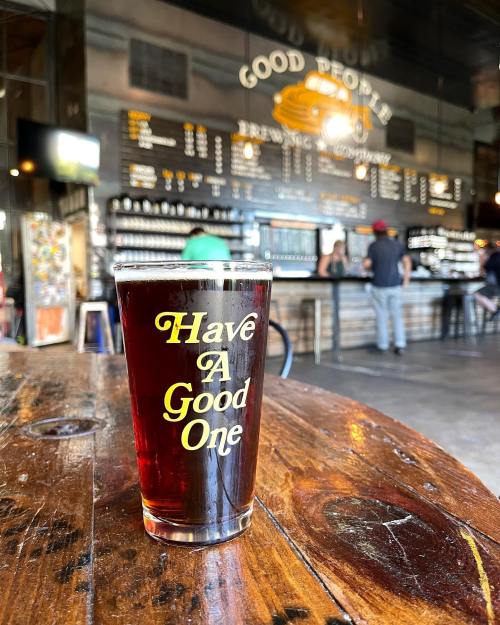 A pint glass with "Have A Good One" sits on a wooden table in a brewery with a bar in the background.