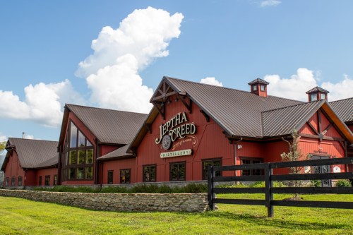 A red barn-style distillery building with a sign reading "Jeptha Creed," set against a blue sky with fluffy clouds.