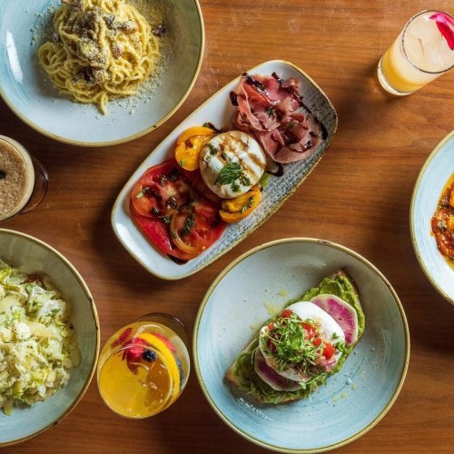 A spread of gourmet dishes including pasta, salad, and colorful appetizers on a wooden table with drinks.