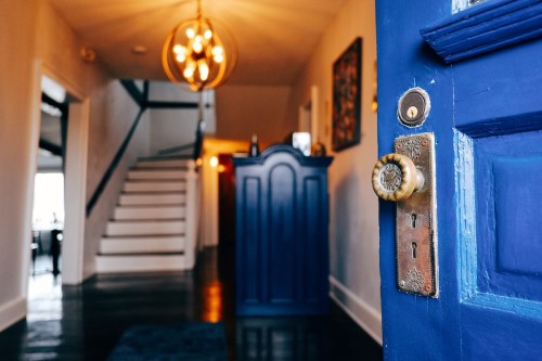 A blue door with a vintage knob opens to a bright hallway featuring a staircase and a stylish light fixture.