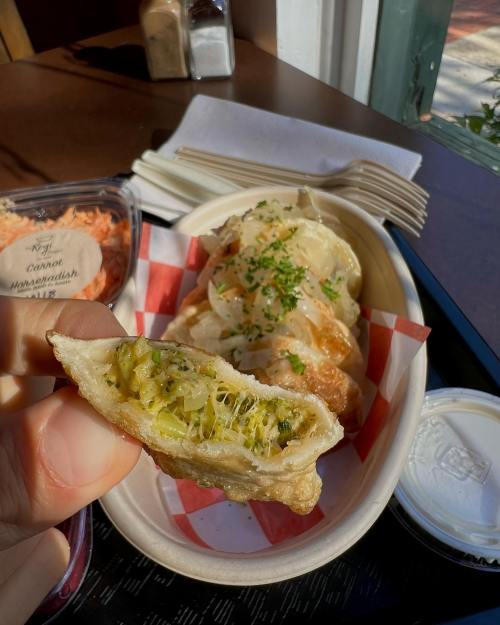 A hand holds a fried dumpling, revealing a green filling, with more dumplings in a basket nearby.