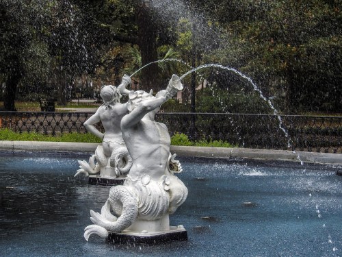 Two white marble mermaid sculptures spouting water in a fountain, surrounded by greenery and a blue pool.