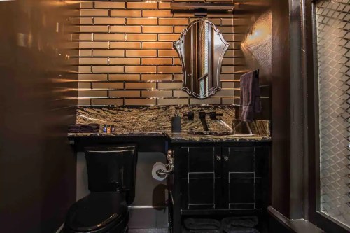 A modern bathroom featuring a black toilet, dark cabinetry, a granite countertop, and a decorative mirror against a brick wall.